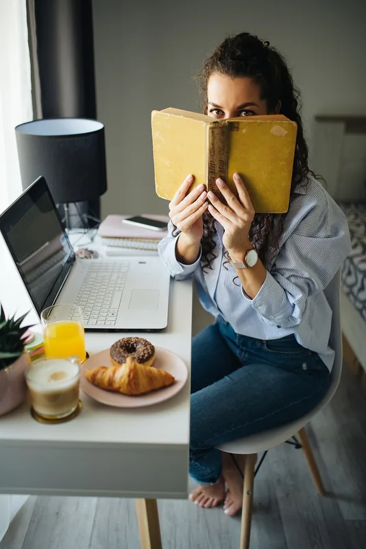 Persona leyendo tranquilamente con un libro y un portátil en un escritorio acogedor de casa, con desayuno y luz natural
